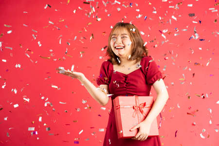 Asian young girl in dress holding red gift box with cheerful expression on backgroundの写真素材