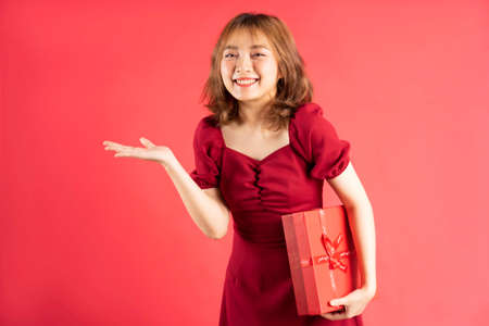 Asian young girl in dress holding red gift box with cheerful expression on backgroundの写真素材