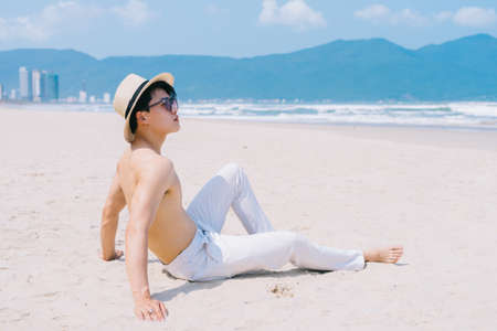 Barebacked young Asian man sitting on the sand and looking at the seaの写真素材