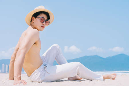 Barebacked young Asian man sitting on the sand and looking at the seaの写真素材