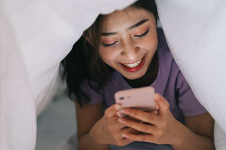 Young Asian girl lying in blanket and using her phone to text with friends on social networksの写真素材