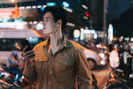 Young Asian man is using his phone while walking in the street at nightの写真素材