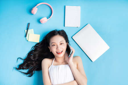 Young Asian girl lying on blue background with laptop, smartphone, headphones and note around