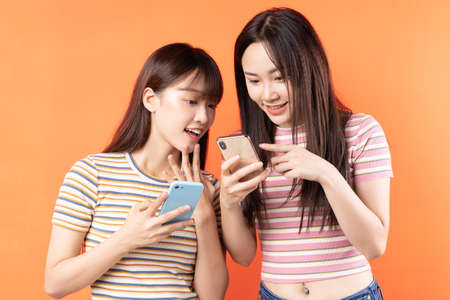 Two beautiful young asian girls are using mobile phones on orange backgroundの写真素材