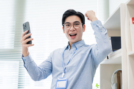 A young man with glasses expresses joy and excitement while holding a smartphone in a bright, modern office. His enthusiastic gesture suggests a recent achievement or good news.の写真素材
