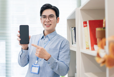 A young Asian man with glasses points at a smartphone while smiling in a contemporary office. The...