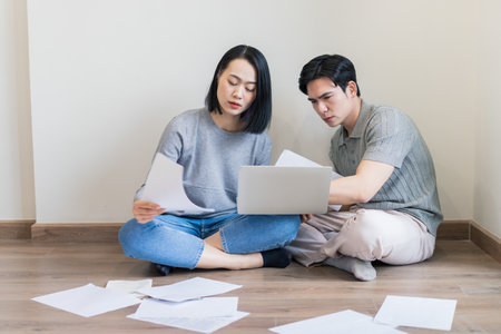 A young couple is seated on the floor, reviewing papers and a laptop. They appear focused and are...