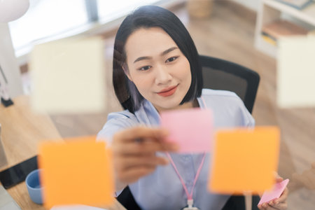 A young Asian woman is focused on arranging colorful post-it notes on a glass board in a contemporary office. She appears engaged and thoughtful while planning her tasks.の写真素材
