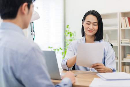 A woman smiles while handing over a document to a man during an interview in a bright office. Their focused expressions reflect a professional atmosphere in a contemporary workspace.の写真素材