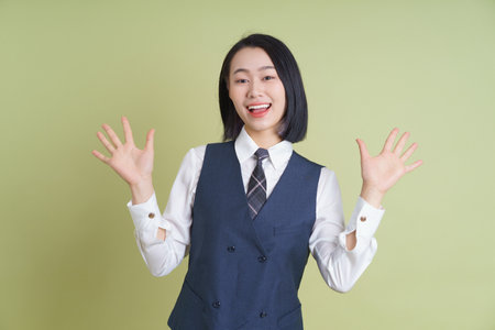 A cheerful young woman dressed in a formal outfit smiles brightly while waving her hands enthusiastically against a light green background. Her outfit and expression convey positivity and excitement.の写真素材