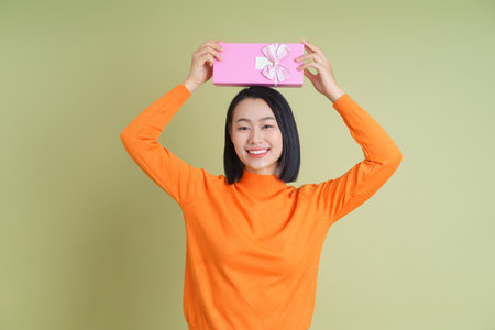 A young Asian woman wearing an orange sweater smiles brightly while holding a pink gift box with a bow above her head. The green background creates a joyful atmosphere.の写真素材