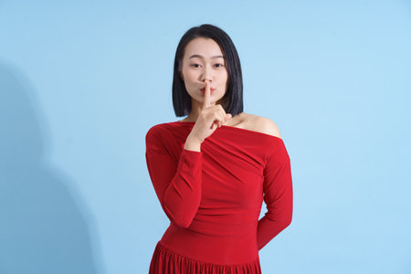 A woman stands against a light blue background wearing a stylish red dress. She gestures silently...