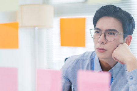 A young Asian man wearing glasses looks thoughtfully at sticky notes during a creative brainstorming...