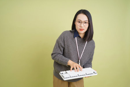 A woman stands in front of a light green background, typing on a white keyboard. She wears glasses and a grey sweater, looking focused and engaged in her activity.の写真素材