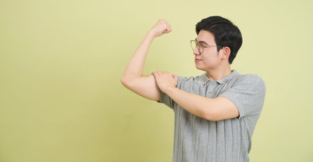 A young Asian man wearing glasses is flexing his arm and displaying his muscle against a light green wall. He appears confident and proud of his strength in this indoor setting.の写真素材