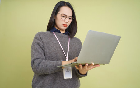 A young Asian woman with glasses engages with her laptop as she focuses intently on her work in a modern setting. She wears a cozy sweater and a lanyard, showing determination.の写真素材