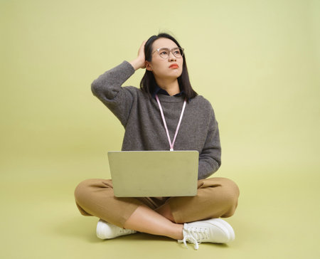 A young woman is sitting cross-legged on a light yellow background, looking contemplative while working on her laptop. She appears to be deep in thought, wearing casual clothing and glasses.の写真素材