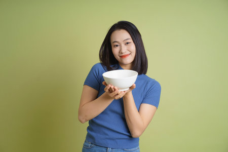 A young woman stands in front of a light green background, smiling while holding a white bowl with both hands, showcasing a warm and friendly demeanor.の写真素材