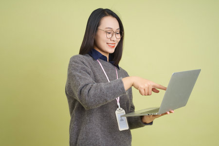 A young Asian woman in a cozy sweater uses her laptop while smiling and pointing at the screen. The...