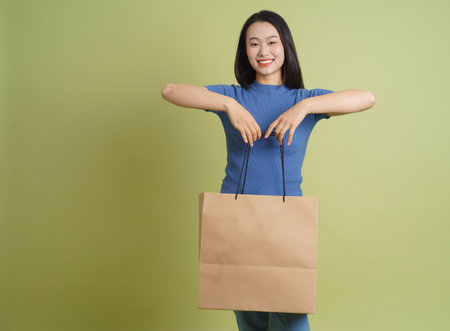 A young Asian woman stands confidently holding a large brown shopping bag. She wears a blue top and expresses happiness, with a pastel green background accentuating her outfit.の写真素材