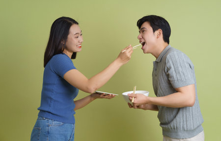 A young couple shares a lighthearted moment as one feeds the other with chopsticks in a charming, green-toned indoor space. Their smiles reflect joy and connection.の写真素材