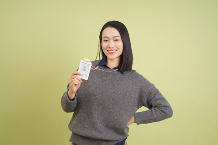 A woman stands proudly holding an identification badge, wearing a cozy sweater. She has a relaxed smile and is positioned in front of a light green wall.の写真素材