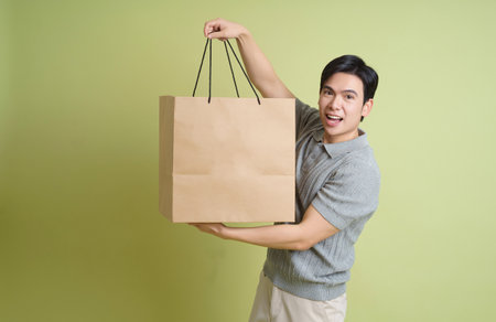 A young man with short black hair joyfully holds up a large brown shopping bag against a vibrant green backdrop, showcasing excitement and enthusiasm.の写真素材