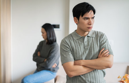 In a bright yet minimalist living room, a man stands with his arms crossed, displaying frustration while a woman sits turned away, both lost in their thoughts, highlighting emotional distance.の写真素材