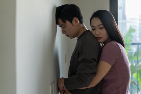 A woman gently embraces a man from behind as he leans against a wall, conveying a moment of vulnerability and closeness in their shared space.の写真素材