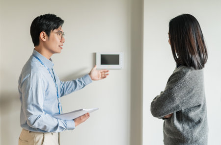 A man presents smart home technology features to a woman in a modern indoor space. The man holds a clipboard, while the woman engages attentively, leaning slightly with crossed arms.の写真素材