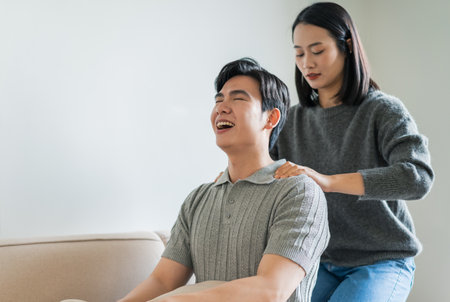 A woman gives a massage to a smiling man in a stylish living room. Natural light filters in, creating a calm environment perfect for relaxation and bonding.の写真素材
