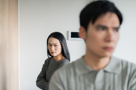 A man stands with his back turned, looking away, while a woman gazes at him disapprovingly. They appear to be in a modern home, sharing an uncomfortable silence.の写真素材
