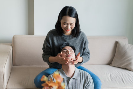 A woman is giving a facial massage to a man while they sit comfortably on a couch in their living room. Both share smiles and laughter, creating a warm atmosphere in the afternoon light.の写真素材