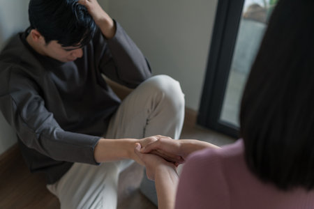 A young man sits on the floor looking distressed while a young woman reaches out to hold his hand gently, offering comfort in a serene indoor space.の写真素材