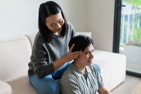 A woman gives a gentle massage to a man while they sit together in a bright, contemporary living room filled with plants during a calm afternoon. Their expressions convey relaxation and connection.の写真素材