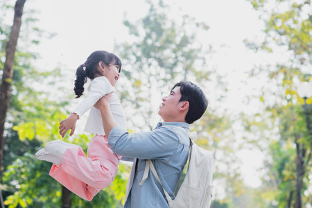 A father delights in lifting his daughter high in the air, both laughing together. They are in a lush park with vibrant trees and bright sunlight, enjoying a warm spring day.の写真素材