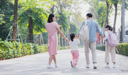 A family strolls through a sunny park in spring, holding hands while two children playfully skip...