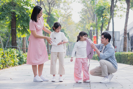 A family of four happily strolls through a park on a bright day. Two children share laughter while their parents assist them, showcasing a warm bond during their outing.の写真素材