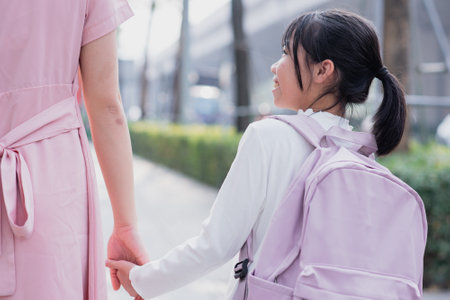 A child dressed in a white top and purple backpack walks hand in hand with an adult, enjoying a...