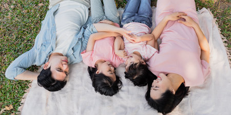 Four family members, including two children, relax on a blanket in a vibrant park. They joyfully interact with each other under the warm sun, creating cherished memories.の写真素材