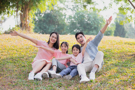 A joyful family of four spends a sunny day in a park, sitting on the grass with arms outstretched in playful poses. The children and parents share laughter and warm moments together.の写真素材