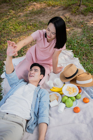 A young couple relaxes on a blanket in a green park. They share a joyful moment while surrounded by a spread of fruits and snacks, enjoying a sunny day together.の写真素材