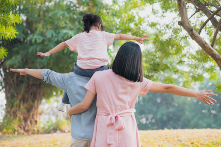 A family spends quality time together in a park. A child sits on the father's shoulders, both arms outstretched in joy. The mother smiles beside them, immersed in a carefree moment.の写真素材