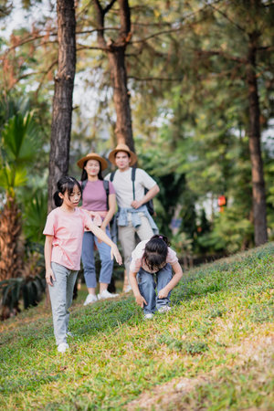 Two children explore a grassy hill in a park, engaging in playful activities under the sun. Family members watch nearby, enjoying each other's company in a vibrant, green environment.の写真素材