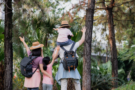 A family explores a vibrant park filled with greenery, enjoying the warm afternoon. Children playfully ride on their parent's shoulders, smiling and raising hands in joy.の写真素材