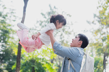 A father lifts his laughing daughter high in the air, creating a joyful moment between them. The park is alive with green trees and bright sunlight, enhancing their fun experience.の写真素材