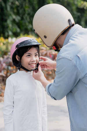 A child wearing a white long-sleeve shirt smiles as a parent fastens a pink and black helmet during a pleasant outdoor day, surrounded by green trees and colorful foliage.の写真素材