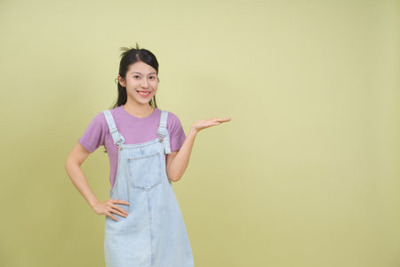A cheerful young woman stands against a solid light green background, wearing a purple shirt and denim overalls. She is smiling and extending her hand outward, invitingly.の写真素材