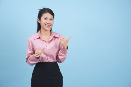 A young woman dressed in a pink blouse and black skirt stands against a blue backdrop. She smiles brightly and gestures with her hands, inviting attention with her cheerful demeanor.の写真素材