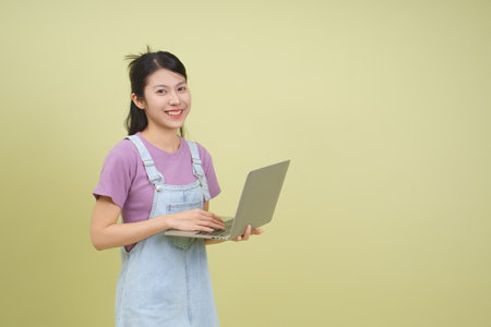 A cheerful young woman stands confidently holding a laptop, showcasing her casual style in a relaxed environment with a soft green backdrop, ideal for remote work or online learning.の写真素材